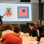 A photograph of a group of adults seated at a long table, taking notes while a person stands at the front of the room, gesturing at a presentation projected on a large screen. The presentation shows two colorful geometric designs.