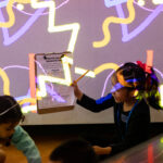 A photograph of two children sitting on the floor. The child on the right is holding up a clipboard and pencil and is looking enthusiastically at the other child. On the wall behind them is a projection of multiple, scattered orange and purple squiggly lines.