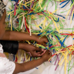 A photograph of a young dark-skinned child with braids, ties a piece of string onto a vertical wire attached to the wall. The wall has multiple pieces of red, blue, yellow, purple, and white coloured string tied to it. There is a larger dark-skinned hand next to the child’s, helping them tie the string.