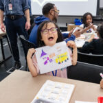 A photograph of a light-skinned child with short straight black hair and purple rimmed glasses smiles enthusiastically holding up a drawing. In the drawing there is a purple and orange builds and a big yellow school bus. In the background there are more young children drawing.