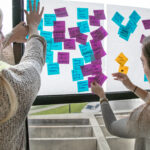 A photograph of two light-skinned adults with long straight hair are placing blue, purple, and yellow sticky notes with text onto a glass window. The text consists of short key words, for example “artist spaces” or “paint cross walks.”