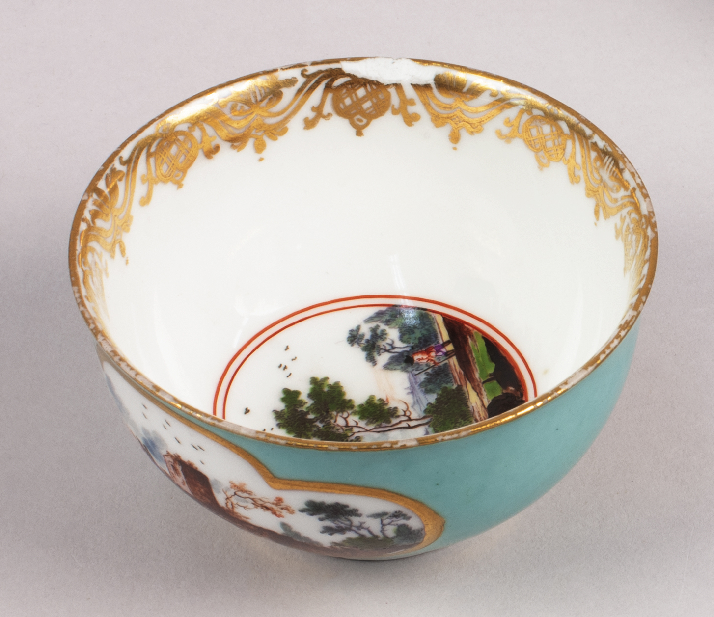 Overhead view of a teacup that is teal on the outside and white on the inside. The rim boasts a decorative gilded pattern and pastoral scenes of featured in vignettes on both the cup’s side and interior bottom.