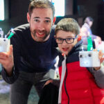 A light skinned adult and child with short brown hair each hold up a white cube that has a face. The cubes have googly eyes, a pointy nose, and a light up mouth. At the top of the cube there are protruding strands of green and white lights, almost looking like hair.