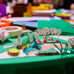 A photograph of a miniature garden constructed out of clay, paper, and cardboard. In the background are scattered craft materials across the table.