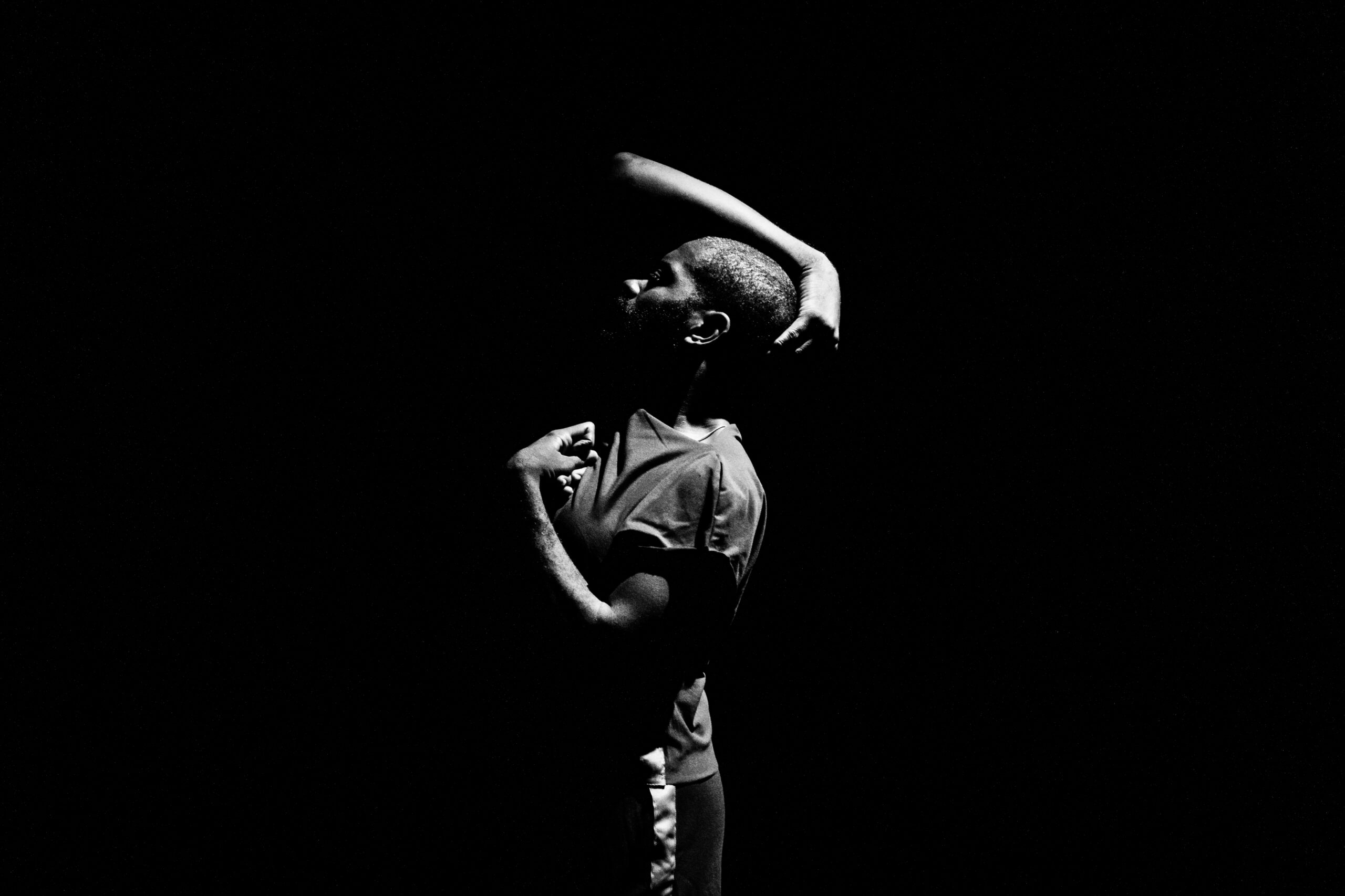 Black and White photo, left side profile of Jerron Herman, a Black man with a Black beard, turns grasping his head in his right arm, while his left arm flexes in a grip by his chest.