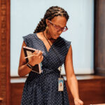 A photograph of a medium-dark skinned adult with long braids, glasses, and wearing a navy polka dot dress holds a note book in one hand and touches a cup sitting on top of a long table. The table is filled with other cups, plates, and a tray.