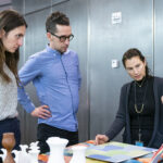 A photograph of a light skinned adult with long brown hair tied into a ponytail, speaks to two adults and is pointing at an object that sits on a small presentation stand. On the table in front of them are vases, and flat colourful patterned artwork. The two adults listening both have their hands on their hips and look deeply concentrated on what the adult is pointing at.