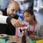 A photograph of a light skinned bald adult and medium skin toned child with long black hair are building a model of a swing set out of pink craft paper. The adult and child look invested in what they are making.