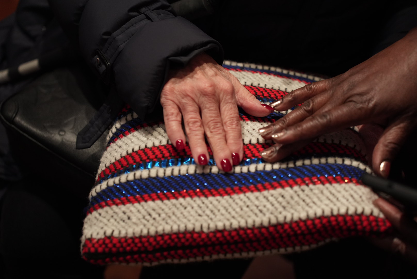 A close up photograph of two hands are feeling the texture of a cushion that has white, red, navy blue, and metallic blue stripes. The hand on the left is light skin toned and has red painted fingernails, and then hand on the right is dark skin toned with shiny copper painted fingernails.