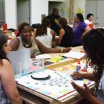 A photograph of three adults sitting around a table scattered with various craft materials such as paper, cardboard, post-it notes, and pencils. The dark skinned adult with long braids is drawing on a constructed model that looks like a garden or outdoor amphitheatre. The dark skinned adult sitting across the table is smiling and looking at what the adult is drawing.