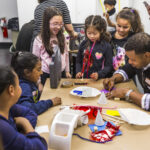 A photograph of a group of children and an adult are gathered around doing a craft activity. There are materials scattered around the table such as pipe cleaners, paper coffee filters, and scissors. The dark-skinned adult is drawing and the kids in the middle designed something made of cardboard, pipe cleaners, and clear colored plastic.