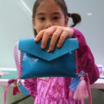 A photograph of a medium light skinned child with dark pigtails and wearing a pink shirt holds up a blue purse close to the camera lens. The blue purse has neon pink stitched sides and pink and blue tassels on bottom corners.