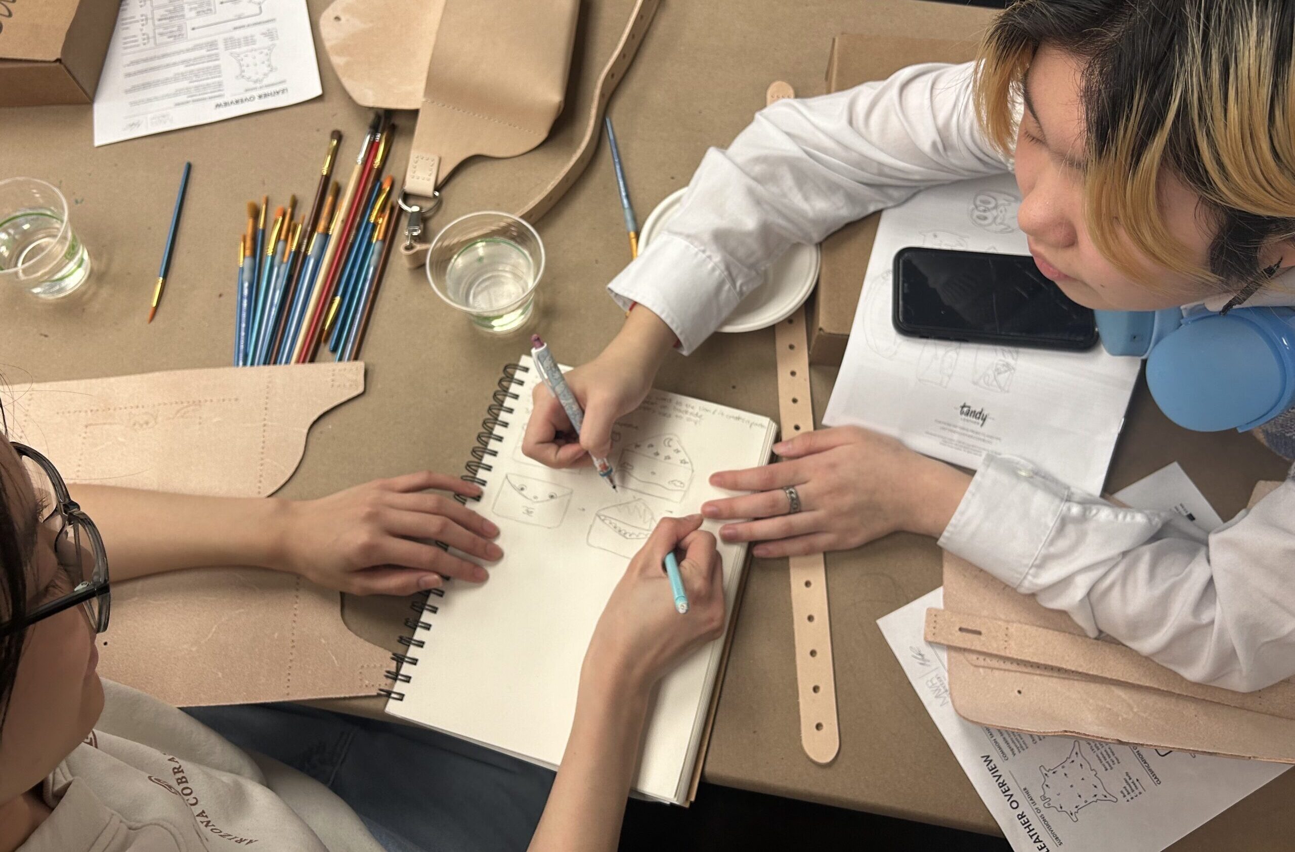A photograph of a bird's eye view of two medium-skin toned teenagers sitting at a table and drawing on a notepad. They are drawing something in the shape of an envelope. Around them are materials such as paint brushes, leather straps and sheets, and worksheets on how to construct a leather accessory.