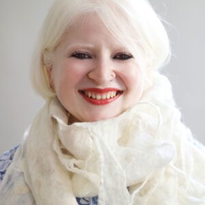 Sugandha sits in front of a neutral background with a bright smile adorned with a simple gold hooped nose ring. Looped around her neck is a pale scarf that matches her pale blonde hair.