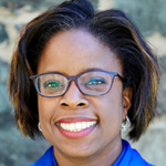 A person with short dark hair smiles. They are wearing dark rimmed glasses and a blue shirt.