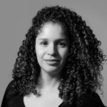 A person with voluminous and curly hair faces forward. They wear a black top. The image is in black and white.