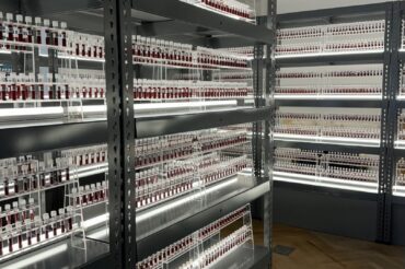Hundreds of vials holding a red substance line brightly lit metal shelves. Each vial has a white stopper and a label.