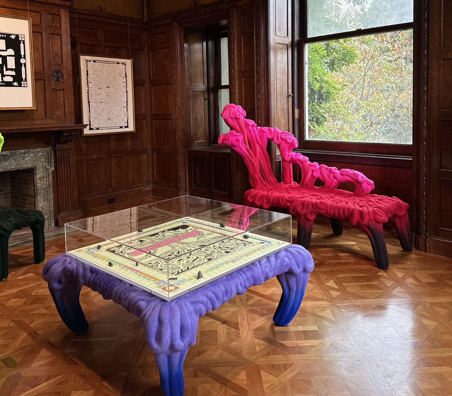 Formed with what appears to be fluorescent-toned, thick cords, a hot pink settee and grape-purple table sit in a room with wood paneling. A square game board fills the top surface of the low table.