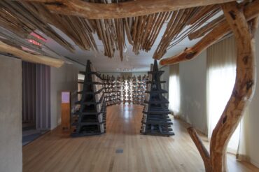 A flat roof made of twigs and branches laid across larger branches, which is held up with a concrete slab to one side and a thicker branch to the other, is above us. Across the room scaffolding with blocks of earth curves in a U shape away from us.