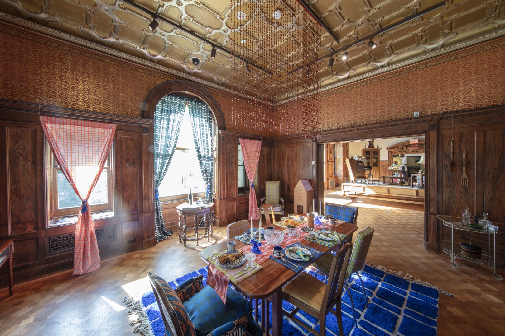 A dining table is set with vividly colored and boldly patterned placemats and napkins. Chairs, settees, the area rug, and curtains have similarly striking designs. The room is paneled in wood beneath gilded wallpaper and a carved ceiling.