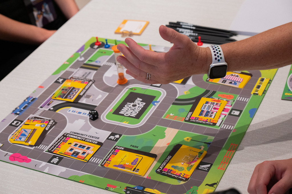 A hand throws a die over a board game titled Design Around Town.
