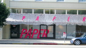 Photograph of a building facade with the word [PAPER] in hot pink capital letters splashed across its awning and windows.