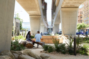 Pedestrians sit on wood benches in a rocky landscaped park underneath the massive concrete pillars of an active overpass.