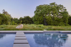 A blue outdoor pool with a stone pathway leading out to a grassy tree-lined landscape decorated with a large rough-hewn boulder.