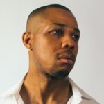 Portrait photo of a Davone Tines with pearl earring and white collard shirt looking to the side.
