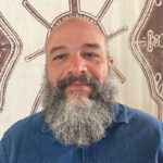 A man with a long beard wearing a blue shirt smiles. The background is a light brown and white geometric patterned tapestry.