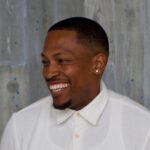 A man in a white collared button-up shirt looks to the left, smiling. The background is light gray with blue specks.