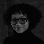 A woman with dark curly hair wearing a black turtleneck and dark framed glasses smiles. The image is in black and white, and the background is dark gray.