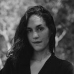 A woman with long dark hair wearing a black blouse. The background is blurred trees and foliage. The photo is in black and white.