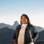 A person with long, brown hair wearing a white high neck shirt and dark jacket stands in front of a mountain landscape.