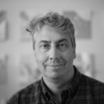 A black and white photo of a person with short hair wearing a dark plaid collared shirt.