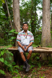 A person wearing a multi-colored striped shirt sits on a wooden bench outdoors. Trees and foliage surround the bench.