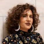 A person with voluminous, curly hair faces forward. They are wearing hoop earrings and a floral top.