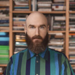 A person with a long beard faces forward. They are wearing a blue, green, and purple striped collared shirt.