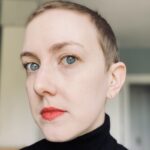 A person with short, blonde hair faces forward. They are wearing a black turtleneck shirt.