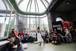 Jerron Herman, a black man in a white cloth dress, dances in the middle of a seated crowd. He reaches his right hand up above his head and leans forward.