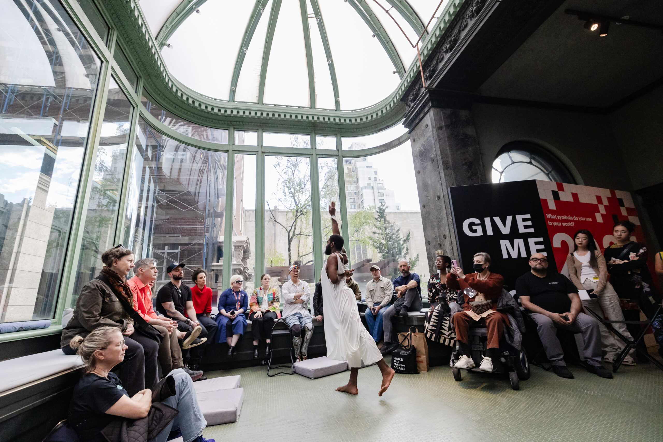 Jerron Herman, a black man in a white cloth dress, dances in the middle of a seated crowd. He reaches his right hand up above his head and leans forward.