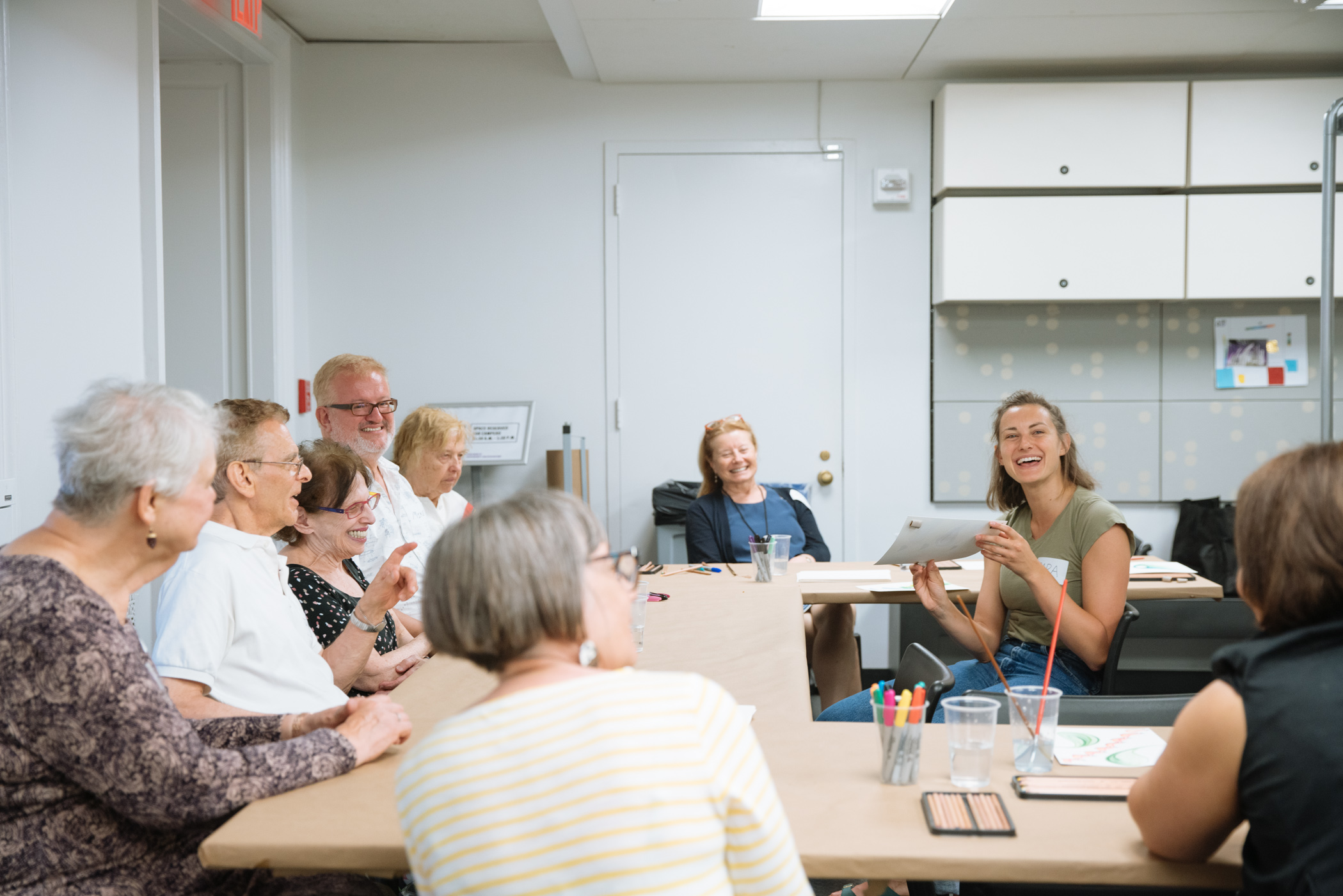 Group of older adults sitting around a U-shaped table. An educator sits in the inside of the U-shaped table showing the group a piece of paper.