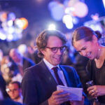 Two guests smile as they review the program, with the colorful lights and excited guests of the National Design Awards just visible in the blurred background.