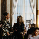 Two women stand to converse at the National Design Awards, with guests seated around them. Behind them, a podium with the Cooper Hewitt logo is framed in the large window.