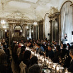 Elegant dinner with guests seated at tables adorned with floral arrangements, listening to Maria Nicanor speak at a podium.