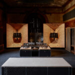 Interior view of a large, high-end audio system, including speakers, subwoofer, and turntable setup. with details in light-colored wood. The system is set up in a room with ornate wood architecture details and a wide window.
