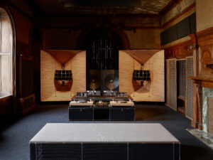 Interior view of a large, high-end audio system, including speakers, subwoofer, and turntable setup. with details in light-colored wood. The system is set up in a room with ornate wood architecture details and a wide window.