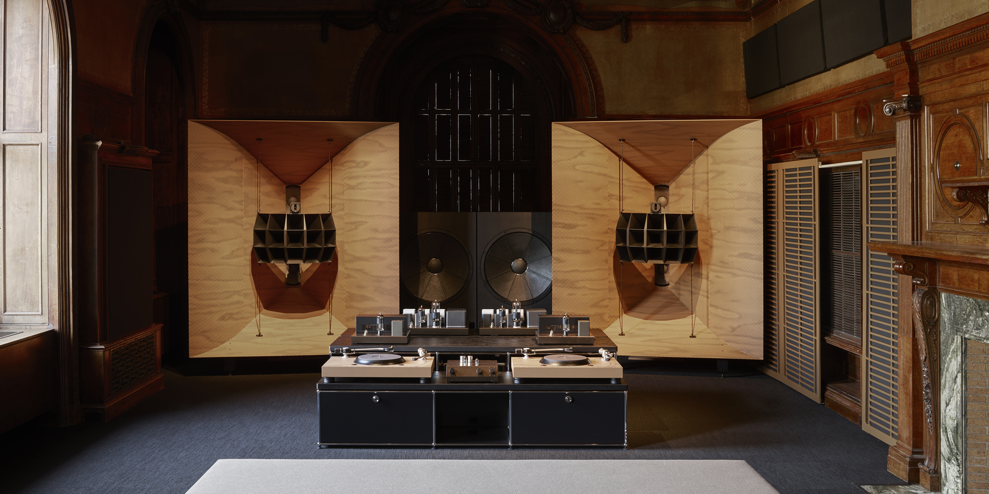 Interior view of a large, high-end audio system, including speakers, subwoofer, and turntable setup. with details in light-colored wood. The system is set up in a room with ornate wood architecture details and a wide window.