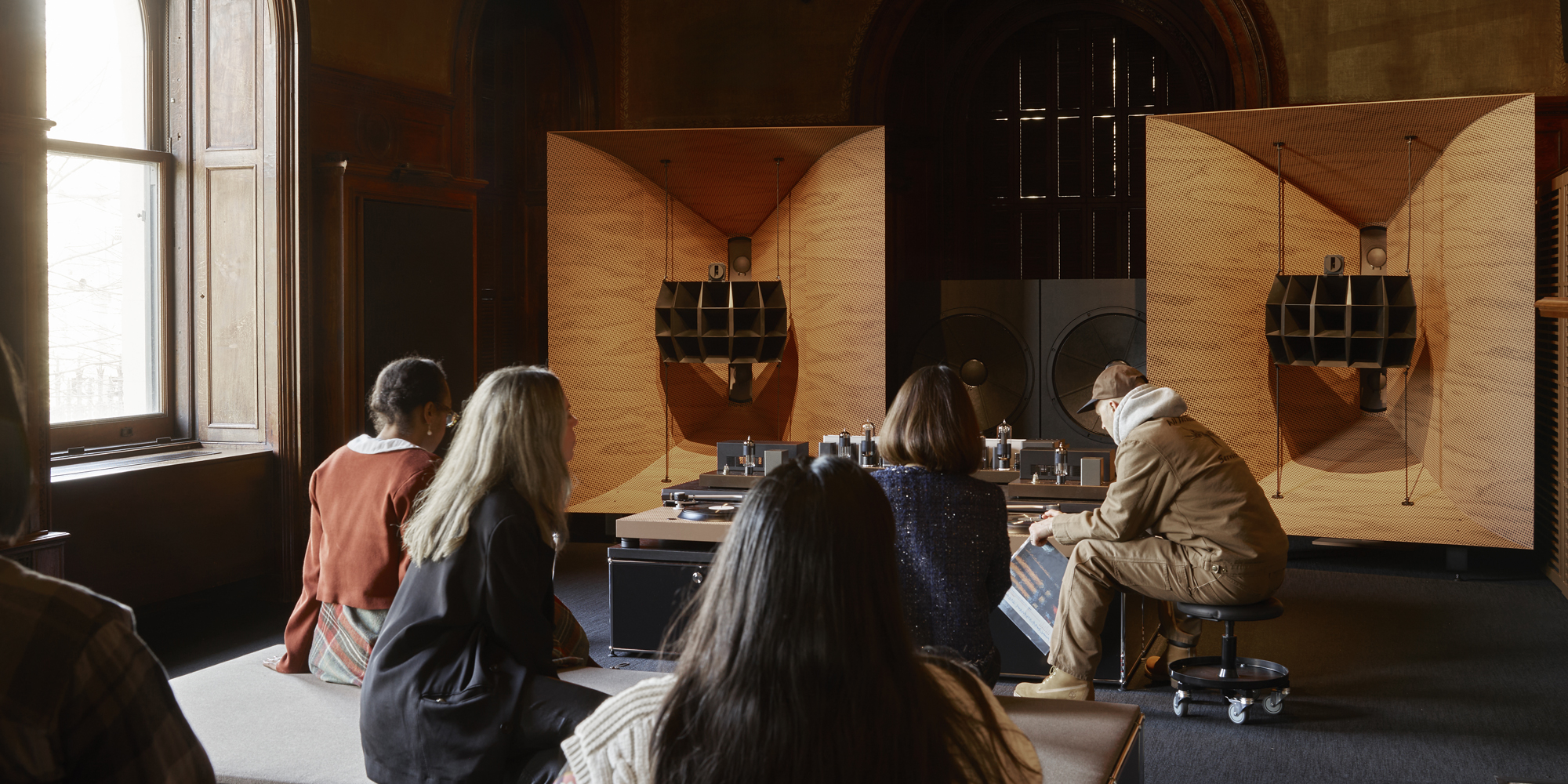 People are seated in front of a large, high-end audio system, including speakers, subwoofer, and turntable setup. with details in light-colored wood. The system is set up in a room with ornate wood architecture details and a wide window.