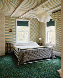 A sunlit bedroom with yellow and green diamond-patterned wallpaper, metal bed frame, and textured green carpet.